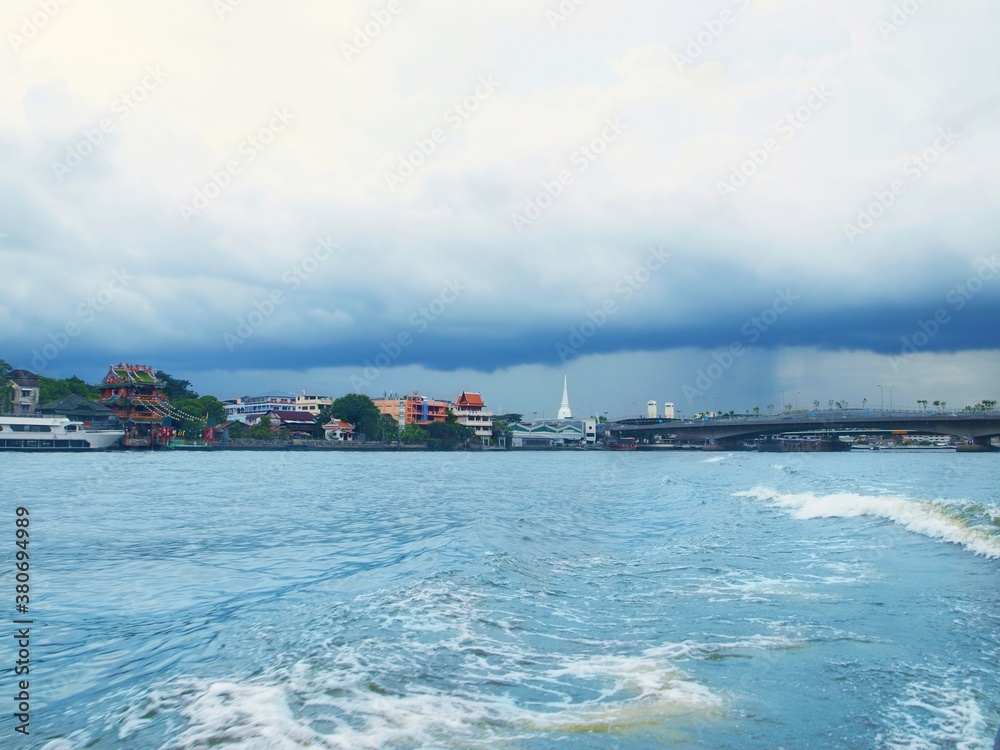 Bangkok, the capital of Thailand, large city. Panoramic view from Chao Phraya River. View of buddhist temples, buildings and bridge in a distance. Blue water, sky with clouds before rain, thundercloud