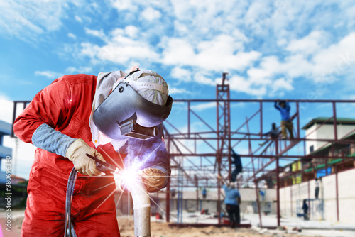 A strong man is a welder in red uniform, welding mask and welders leathers, a metal product is welded with a Tig welding machine at the construction site
