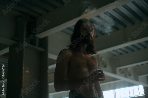 a man with a naked torso smokes in the dark in the parking lot and looks at the camera