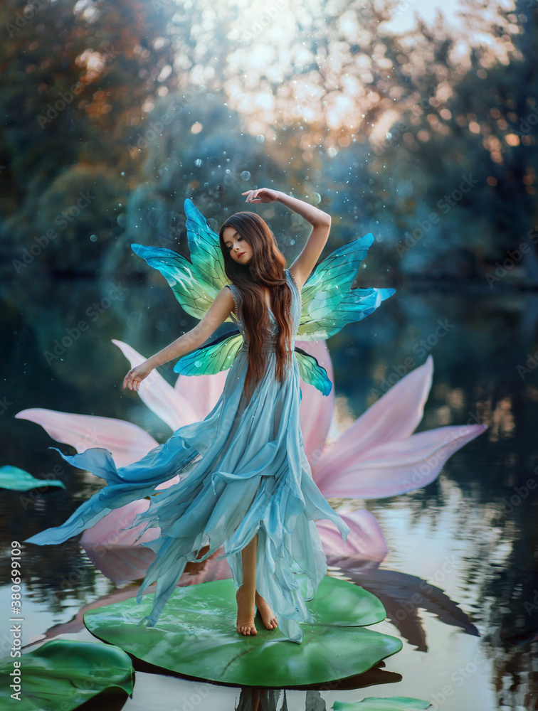 Fototapeta premium Beautiful young fantasy woman in the image of a river fairy dances on a water lily flower. A long silk dress flies in the wind, butterfly wings glisten. Background evening dark nature, blue lake.