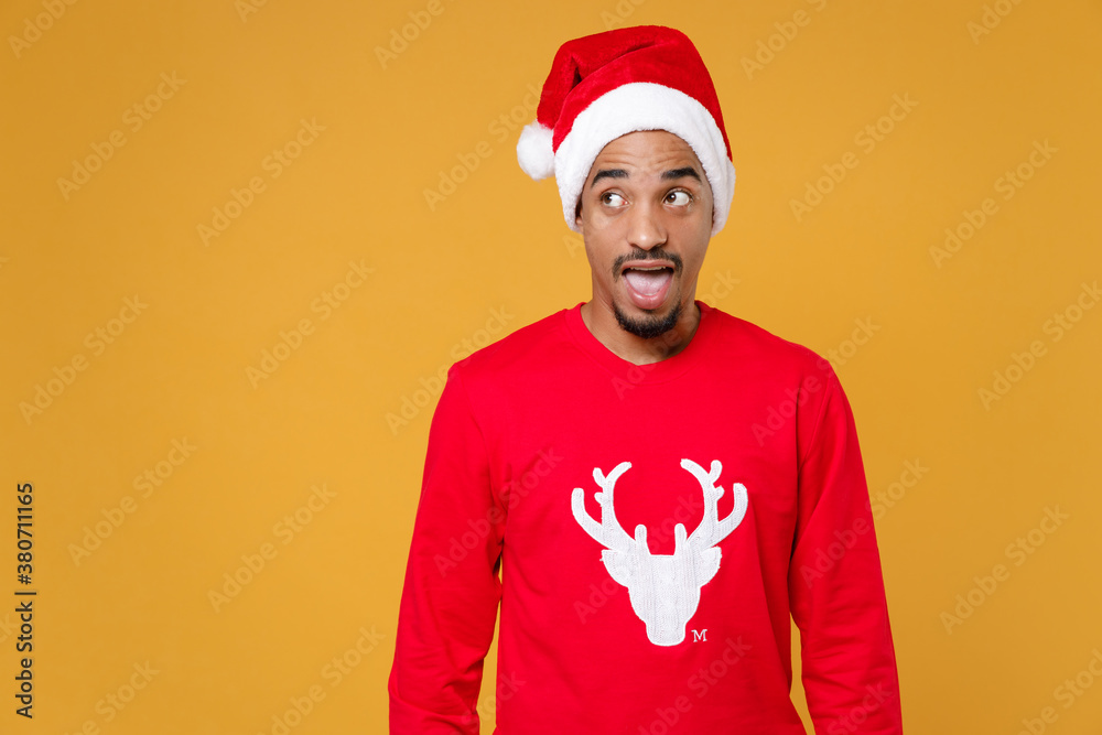 Shocked young Santa african american man in red sweater Christmas hat keeping mouth open looking aside isolated on yellow background studio portrait. Happy New Year celebration merry holiday concept. Shocked young Santa african american man in red sweater Christmas hat keeping mouth open looking aside isolated on yellow background studio portrait. Happy New Year celebration merry holiday concept.