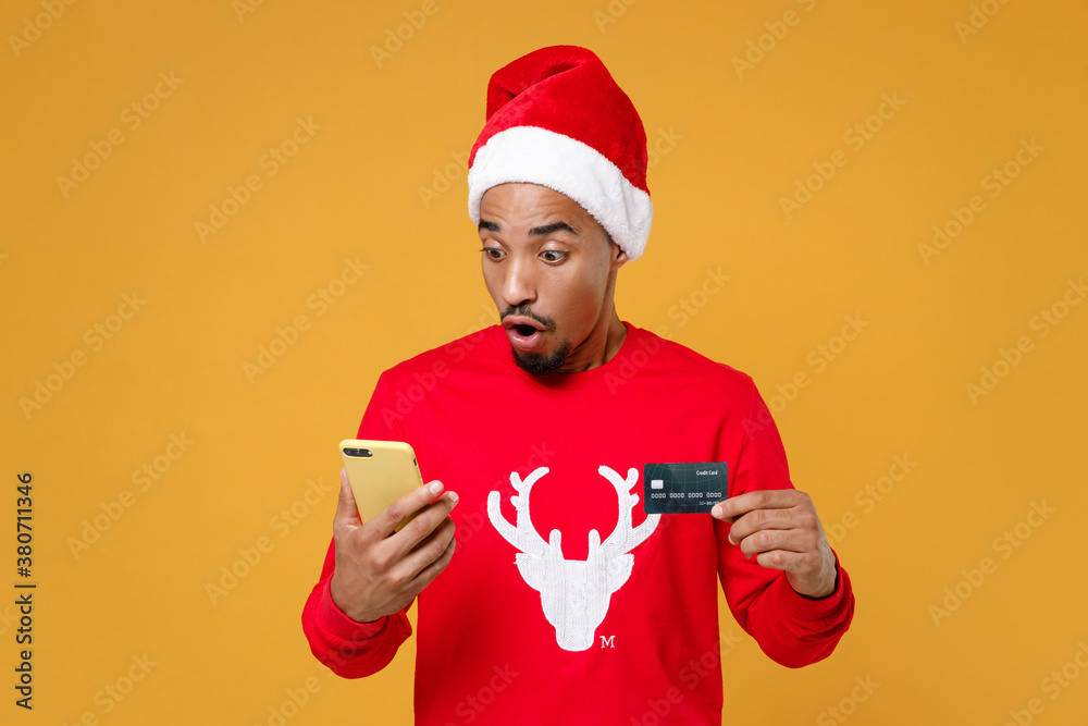 Shocked young Santa african american man in red sweater Christmas hat using mobile cell phone hold credit bank card isolated on yellow background studio portrait. New Year celebration holiday concept.