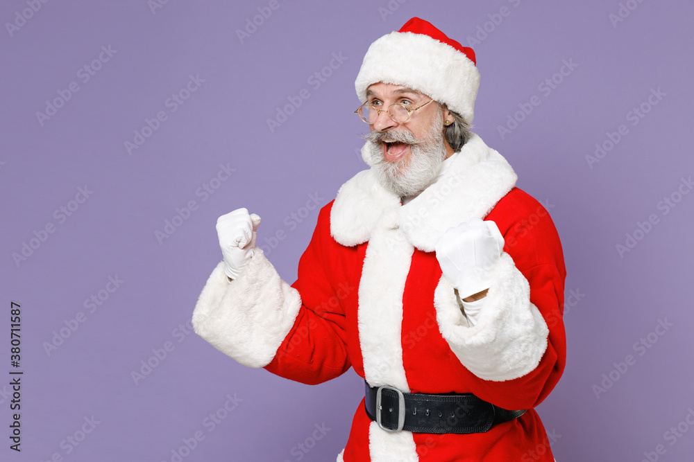 Shocked Santa Claus man in Christmas hat red suit coat white gloves glasses clenching fists doing winner gesture isolated on violet background studio. Happy New Year celebration merry holiday concept. Shocked Santa Claus man in Christmas hat red suit coat white gloves glasses clenching fists doing winner gesture isolated on violet background studio. Happy New Year celebration merry holiday concept.