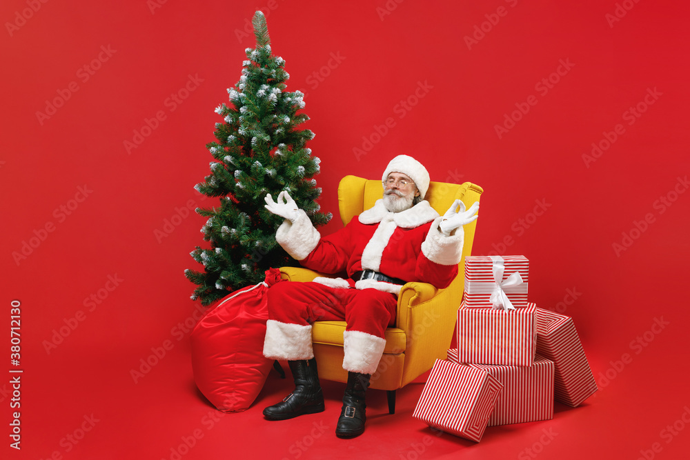 Santa Claus man in Christmas hat suit sit in armchair with fir tree presents gifts hold hands in yoga gesture meditating isolated on red background. Happy New Year celebration merry holiday concept.