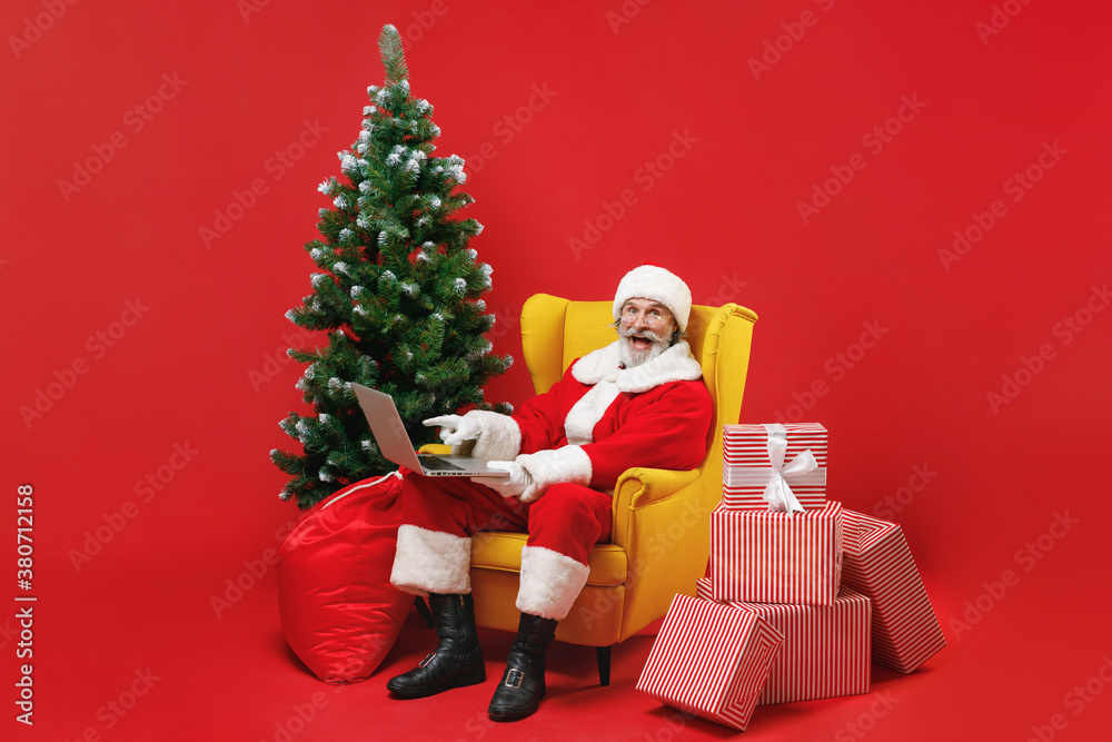 Excited Santa Claus man in Christmas hat suit sit in armchair with fir tree present gifts boxes pointing on laptop pc computer isolated on red background. Happy New Year celebration holiday concept. Excited Santa Claus man in Christmas hat suit sit in armchair with fir tree present gifts boxes pointing on laptop pc computer isolated on red background. Happy New Year celebration holiday concept.