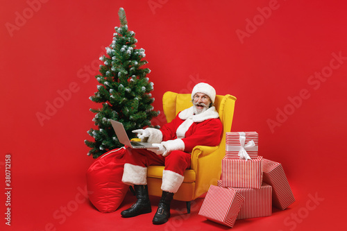 Excited Santa Claus man in Christmas hat suit sit in armchair with fir tree present gifts boxes pointing on laptop pc computer isolated on red background. Happy New Year celebration holiday concept.