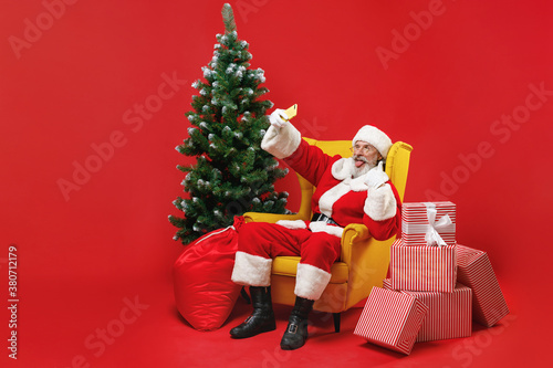 Funny Santa Claus man in Christmas suit sit in armchair with fir tree gifts doing selfie shot on mobile phone showing thumb up isolated on red background. Happy New Year celebration holiday concept.