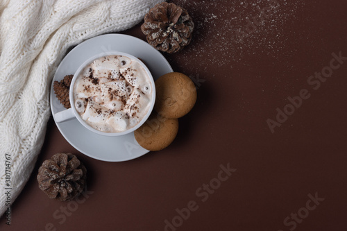 Autumn and winter concept. Top view of cup of cocoa with marshmallows, cookies, white sweater, pine cones and snow on brown background. Chestnut color. Hygge