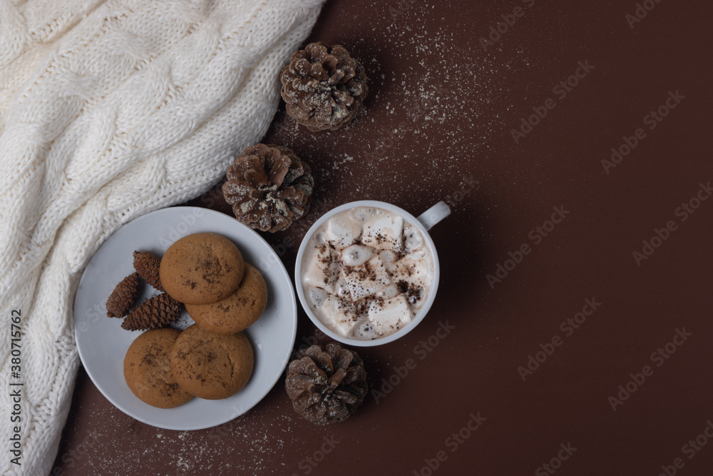 Top view of cup of cocoa with marshmallows, cookies, white sweater, pine cones and snow on brown background. Autumn and winter concept. Chestnut color. Top view of cup of cocoa with marshmallows, cookies, white sweater, pine cones and snow on brown background. Autumn and winter concept. Chestnut color.