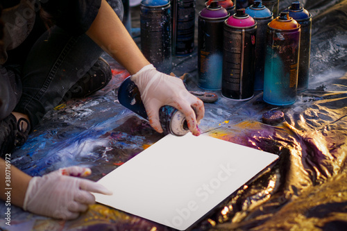 girl street artist in gloves paints with spray cans of paint on a piece of paper. multicolored cans of paint stand around. copy space. place on image