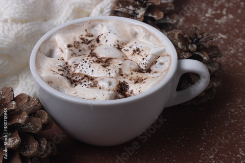 Fototapeta Naklejka Na Ścianę i Meble -  Autumn and winter concept. Cup of cocoa with marshmallows, white sweater, pine cones and snow on brown background. Chestnut color.