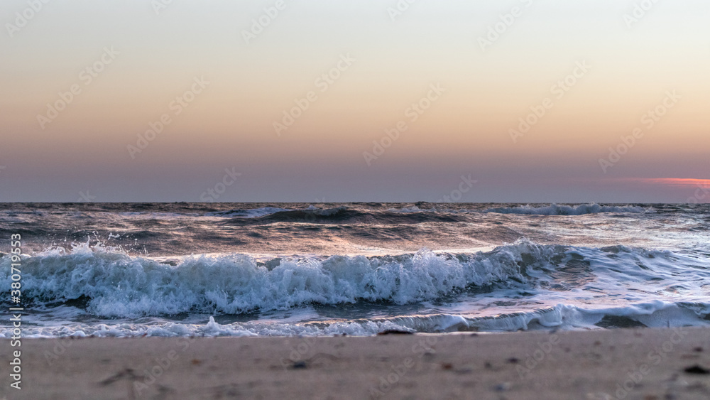 Stormy white glossy waves on sea shore sand beach close-up with sunset sky in pastel colors