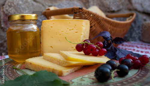 Slices of hard cheese on a ceramic plate, a jar of honey, currant berries on the background of a stone wall