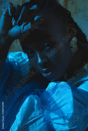 Closeup portrait of a black girl with golden earring and an arm near her forehead sitting in a blue light