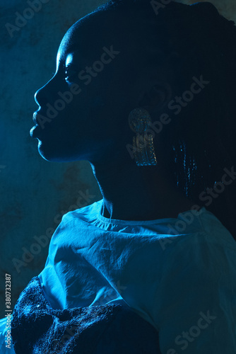 Closeup profile portrait of a black girl with golden earring sitting in a blue light in a fashionable outfit