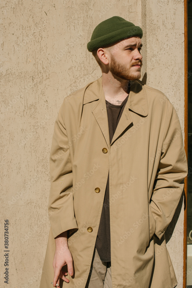 Stylish guy with the read beard in the sand colored trench coat and olive  hat smokes on the street Stock Photo | Adobe Stock