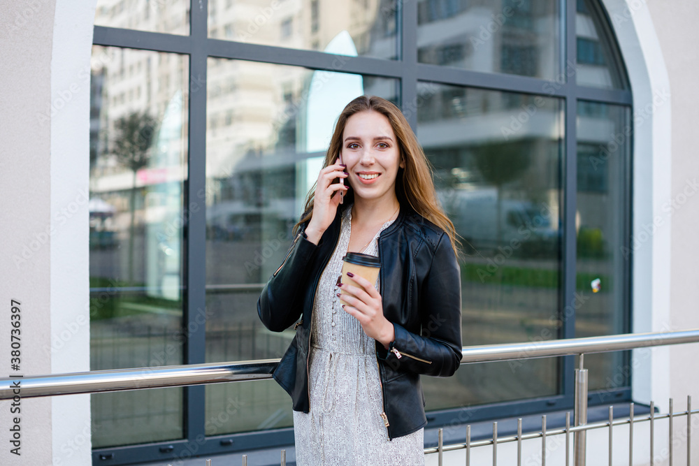 Beautiful woman is speaking on the phone and smiling, with a cup of coffee in the hand, against the glass facade in the city