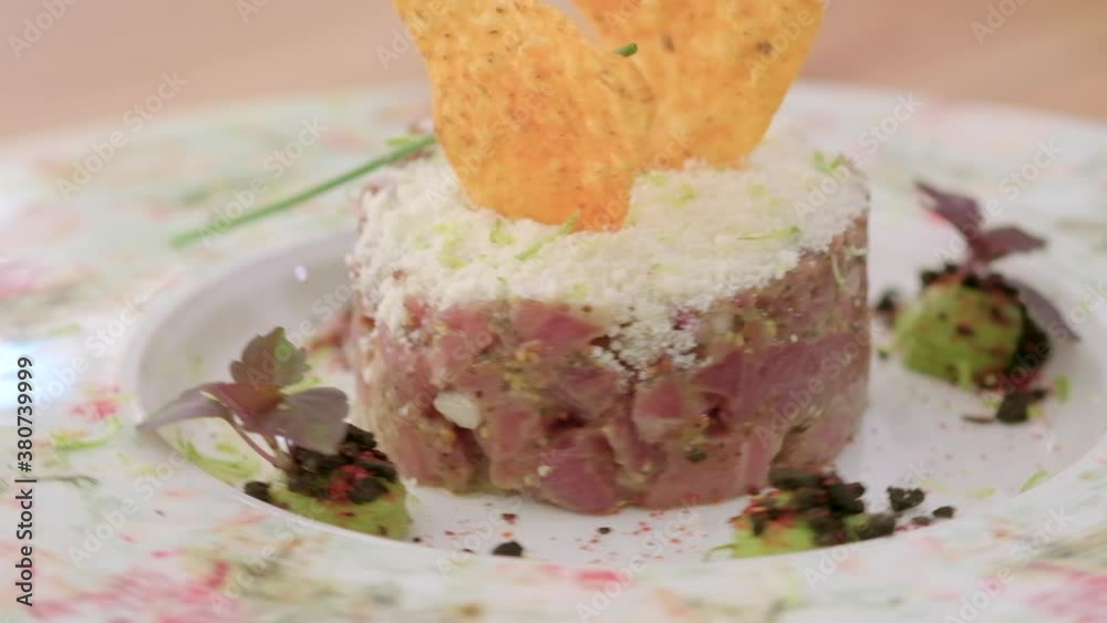 Close up slow motion view at spinning plate served portion of beef steak tartare or tar tar decorated with avocado, parmesan cheese, leaves of salad and chips. Restaurant menu dish concept Close up slow motion view at spinning plate served portion of beef steak tartare or tar tar decorated with avocado, parmesan cheese, leaves of salad and chips. Restaurant menu dish concept