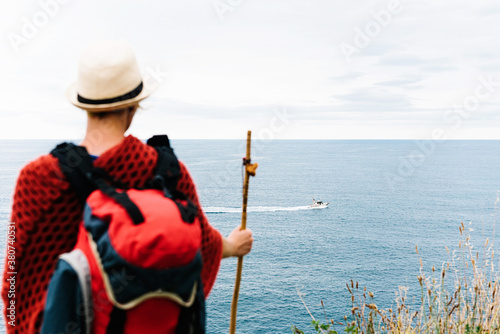 Back view of anonymous female hiker with backpack and stick standing on hill and admiring rippling sea with floating boat while resting during Camino de Santiago route in Spain photo