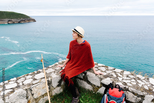 From above female backpacker in stylish clothes sitting on stone border against sea bay and looking away while resting during Camino de Santiago route in Spain photo