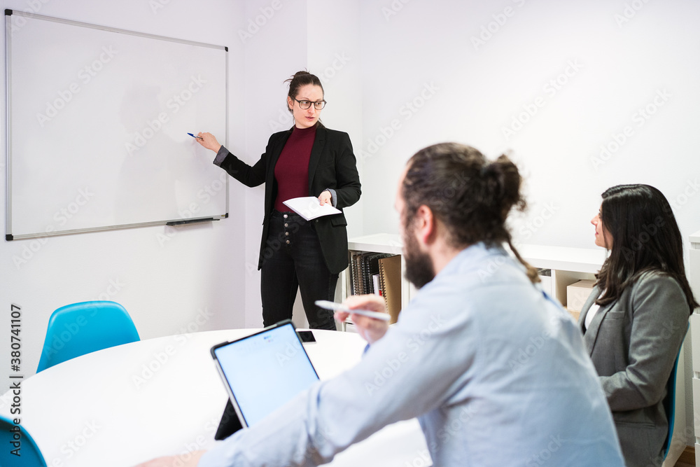 © Alvaro Sanchez/ADDICTIVE STOCK - Young female manager standing near whiteboard and explaining marketing strategy of business project to focused colleagues during presentation in contemporary coworking room