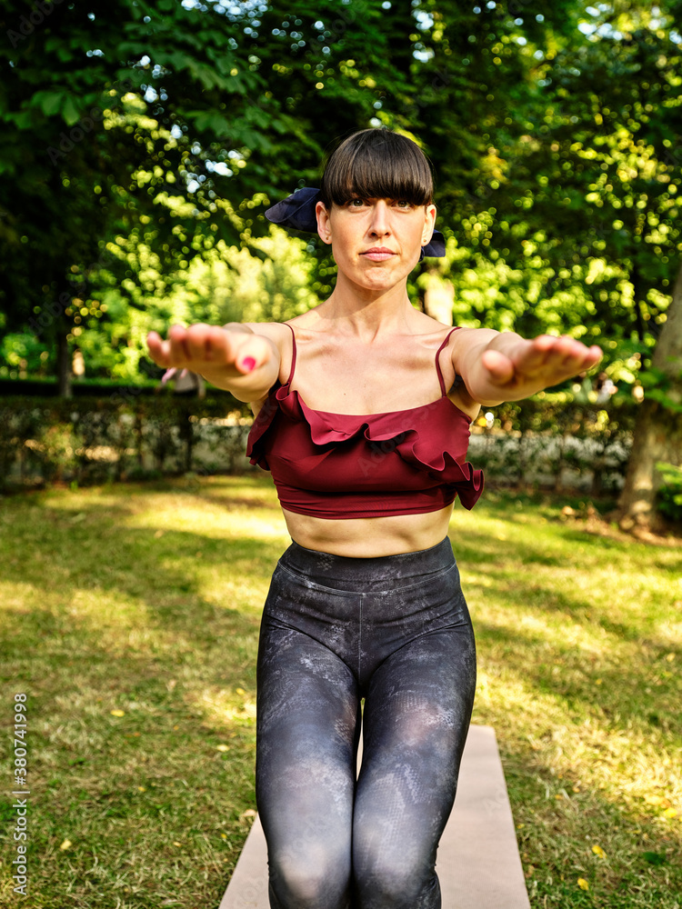 Concentrated slim female in sportswear balancing barefoot on mats in Utkatasana while practicing yoga in green garden looking at camera Concentrated slim female in sportswear balancing barefoot on mats in Utkatasana while practicing yoga in green garden looking at camera