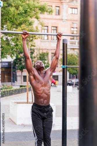 Focused African American male athlete doing pull ups on crossbar during intense training on sports ground in city
