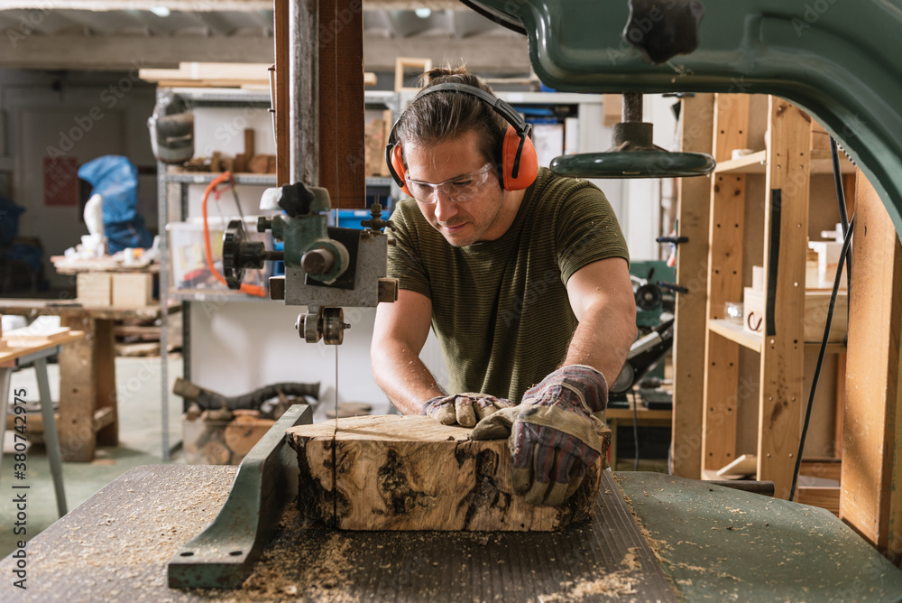 Focused male carpenter in protective ear muffs and goggles cutting piece of wood with band saw in shabby workshop Focused male carpenter in protective ear muffs and goggles cutting piece of wood with band saw in shabby workshop