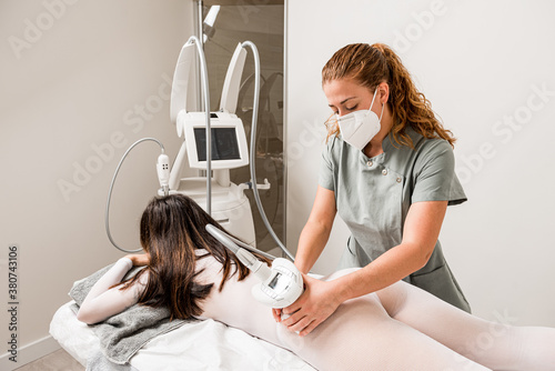 Masseuse in protective mask doing anti cellulite lipo massage to female client buttocks dressed in white suit during body care procedure in salon of apparatus cosmetology