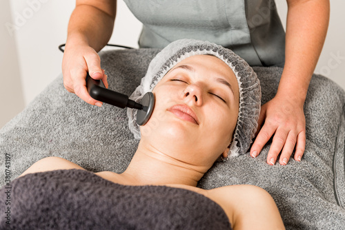 Relaxed female client getting skincare facial massage with ultrasound device from cropped unrecognizable professional cosmetician in contemporary beauty clinic photo