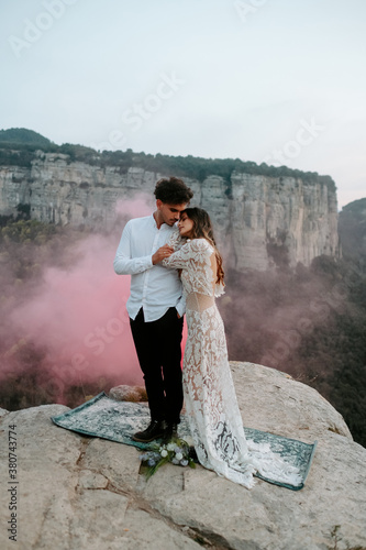 Full body of gentle young couple n stylish wedding outfits standing together with eyes closed on old carpet placed on top of rock with red smoke bomb against amazing scenery of Morro de Labella in Spain photo