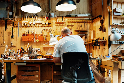 Back view of unrecognizable male luthier master sitting on chair and catching tool from wall while making instruments in workshop photo