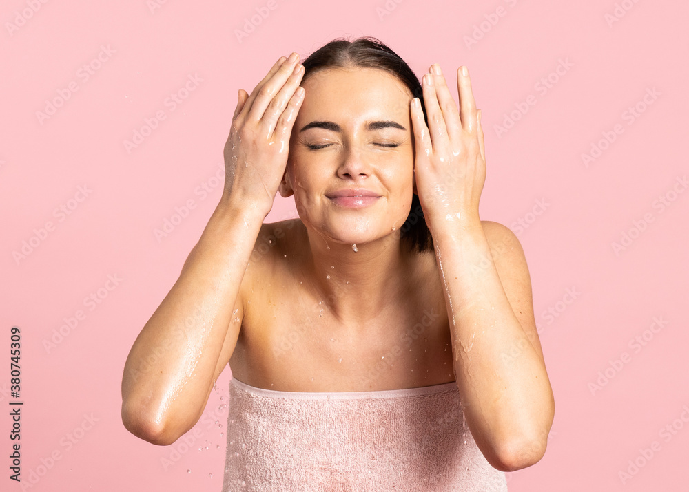 Woman wrapped in towel throwing herself water in the face with closed eyes while standing against pink background Woman wrapped in towel throwing herself water in the face with closed eyes while standing against pink background