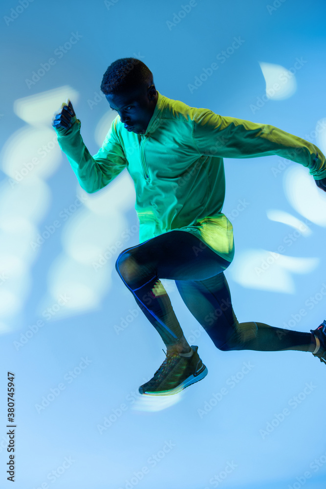 Full body side view of young African American male runner in colorful tracksuit sprinting from start position in studio with bright neon illumination Full body side view of young African American male runner in colorful tracksuit sprinting from start position in studio with bright neon illumination