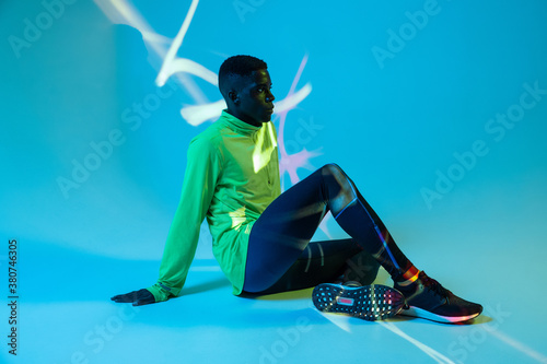 Full body side view of strong young African American male athlete in stylish colorful tracksuit and sneakers sitting on floor in blue studio with neon lights photo