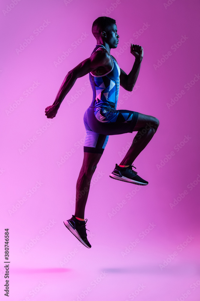 Full body side view of energetic young African American male sprinter jumping against purple neon background Full body side view of energetic young African American male sprinter jumping against purple neon background