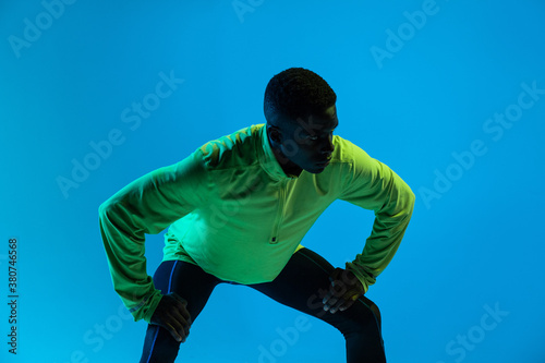 Strong young African American male athlete in stylish colorful tracksuit and sneakers standing in blue studio photo