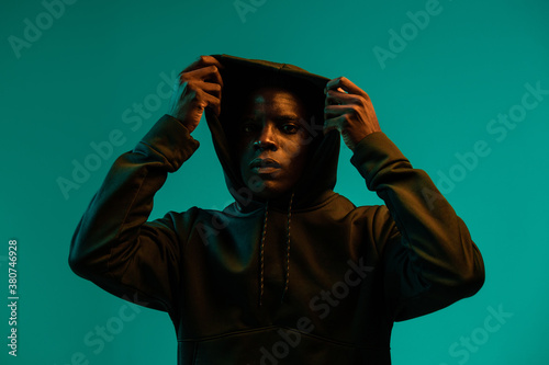 Serious young African American male in black hoodie covering head with hood against green background in studio photo