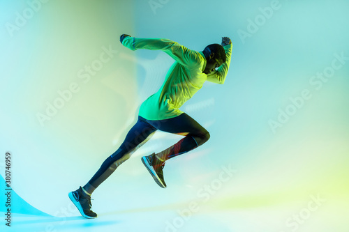 Full body side view of young African American male runner in colorful tracksuit sprinting from start position in studio with bright neon illumination photo