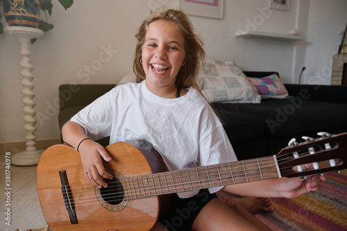 Smiling blonde girl looking at camera while tuning a guitar in the dining room of a house with a sofa and plants