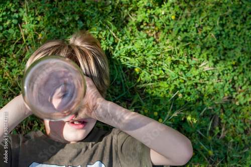 From above of adorable child lying on grass in summer and looking at camera through bottle pretending playing with spyglass