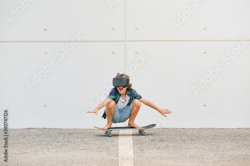 Boy in summer clothes skating on skateboard wearing virtual reality glasses and gesturing with the arms raised on the street
