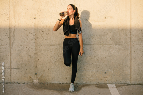 Full body young slim female jogger in black sportswear with towel on neck leaning against concrete wall and drinking nutritive beverage after training on street photo
