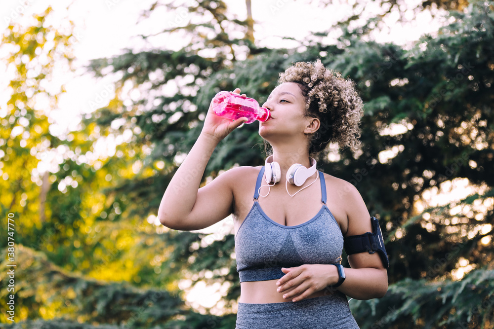 Low angle of young athletic ethnic female with headphones and smartphone drinking refreshing water from plastic bottle while standing against green trees and relaxing after training in park Low angle of young athletic ethnic female with headphones and smartphone drinking refreshing water from plastic bottle while standing against green trees and relaxing after training in park