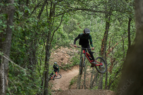 Male cyclists in protective helmets and costumes performing dangerous stunts on bikes for downhill on trail in forest