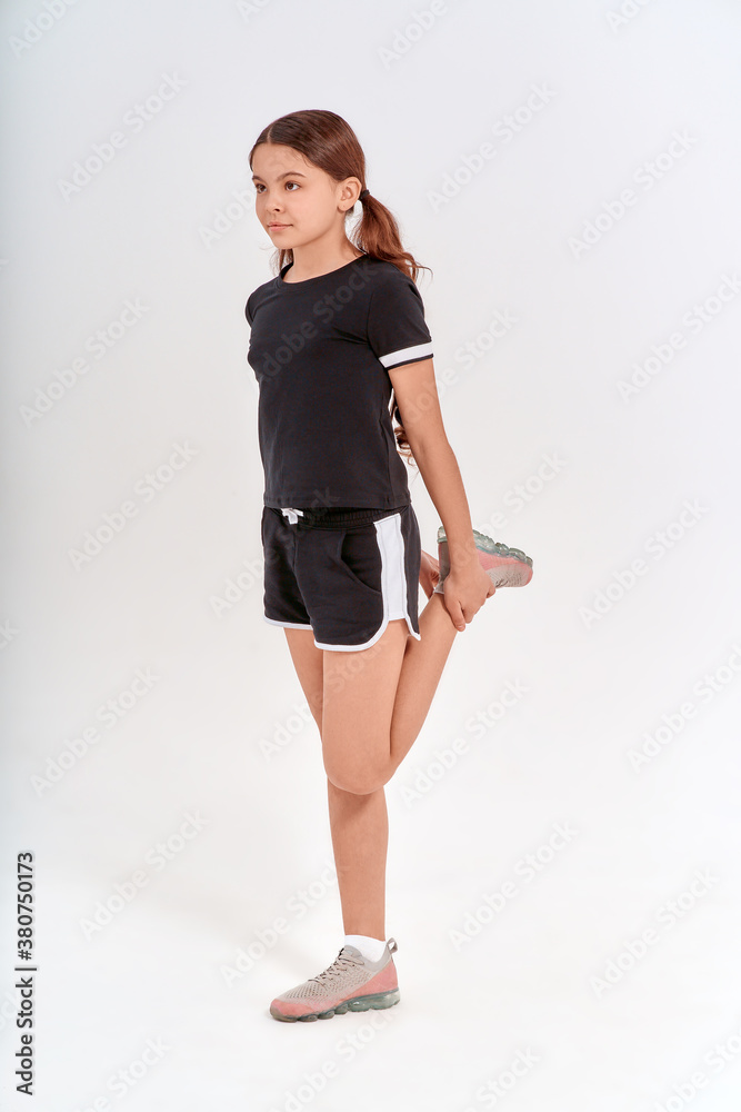 Fototapeta premium Gymnastic exercises. Full length of cute teenage girl in sportswear stretching her legs, warming up while standing isolated over grey background in studio