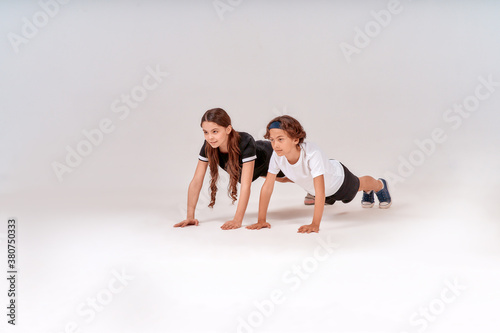 Full-length shot of active teenage boy and girl doing push-ups isolated over grey background, children engaged in sport.