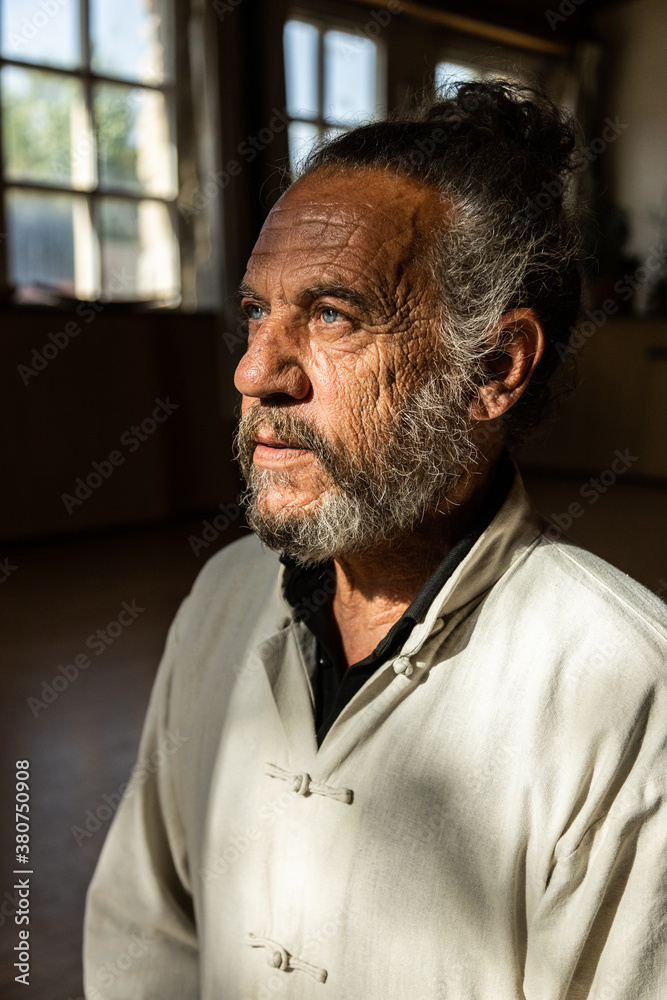 Serious elderly bearded ethnic male instructor of traditional chi kung or qigong looking away while standing in dark room