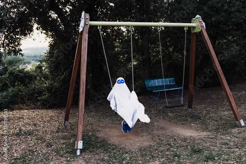 Unrecognizable kid in white ghost costume swinging and playing on playground during Halloween holiday