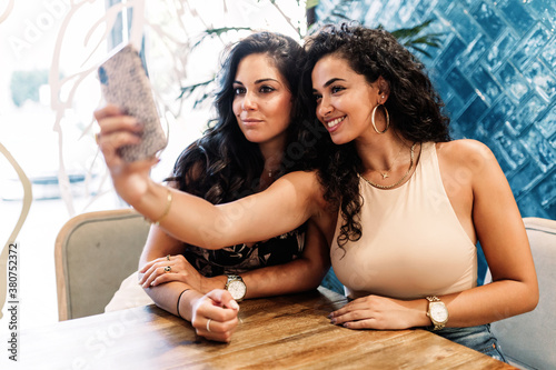 Charming ethnic female friends sitting on terrace of outdoors cafe and taking photo on smartphone